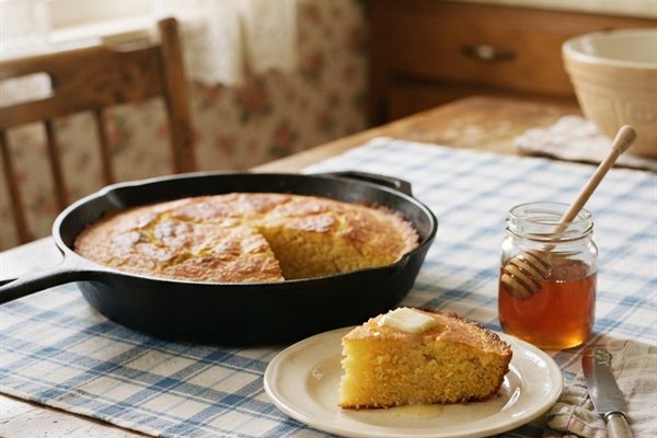 Golden cornbread in a black cast iron skillet on a blue checkered cloth, one wedge on a plate with melting butter and a jar of honey.